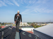 Ein Handwerker mit Helm und Auffanggurt steht gesichert auf einem Laufrost auf einem Ziegeldach.