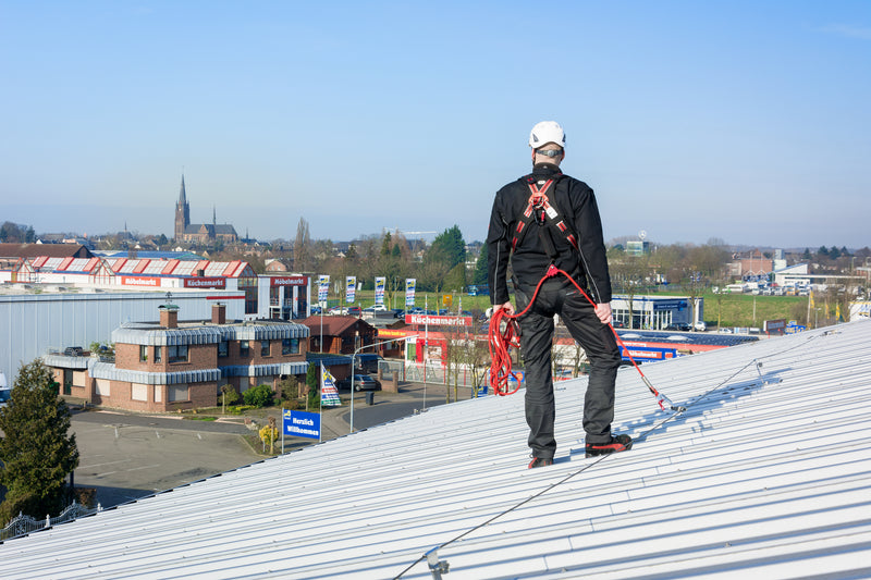 Ein Arbeiter in Schutzausrüstung steht gesichert mit einem roten Seil und Auffanggurt auf einem geneigten Metalldach.