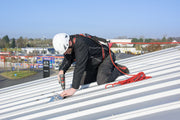 Handwerker mit Helm und Auffanggurt montiert einen Anschlagpunkt auf einem Metalldach mit einem Akkuschrauber.