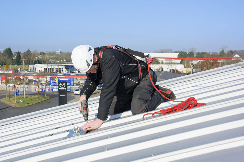 Handwerker mit Helm und Auffanggurt montiert einen Anschlagpunkt auf einem Metalldach mit einem Akkuschrauber.