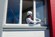 Eine Person reinigt ein Fenster von innen und ist dabei mit einem Auffanggurt und Helm gesichert.