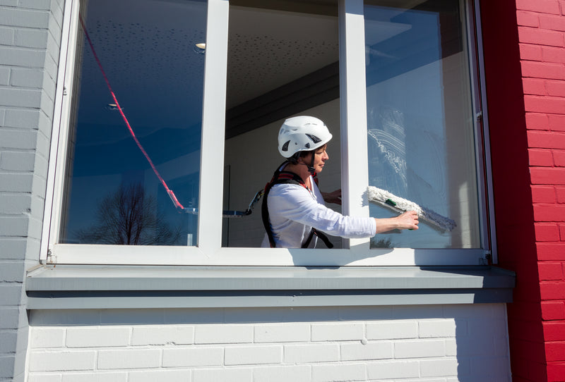 Eine Person reinigt ein Fenster von innen und ist dabei mit einem Auffanggurt und Helm gesichert.