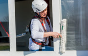 Eine Frau reinigt ein Fenster und trägt dabei einen Auffanggurt mit Verbindungsmittel sowie einen weißen Schutzhelm.