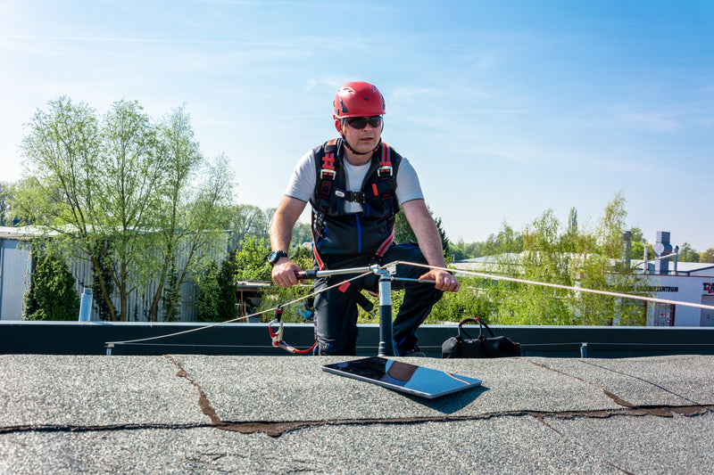 Ein Arbeiter mit rotem Helm und Auffanggurt sichert sich an einem horizontalen Seilsicherungssystem auf einem Flachdach.