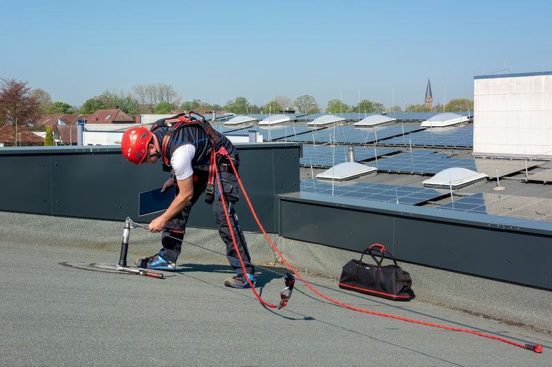 Ein Handwerker mit rotem Helm und Auffanggurt montiert einen Anschlagpunkt auf einem Flachdach mit Solaranlagen.