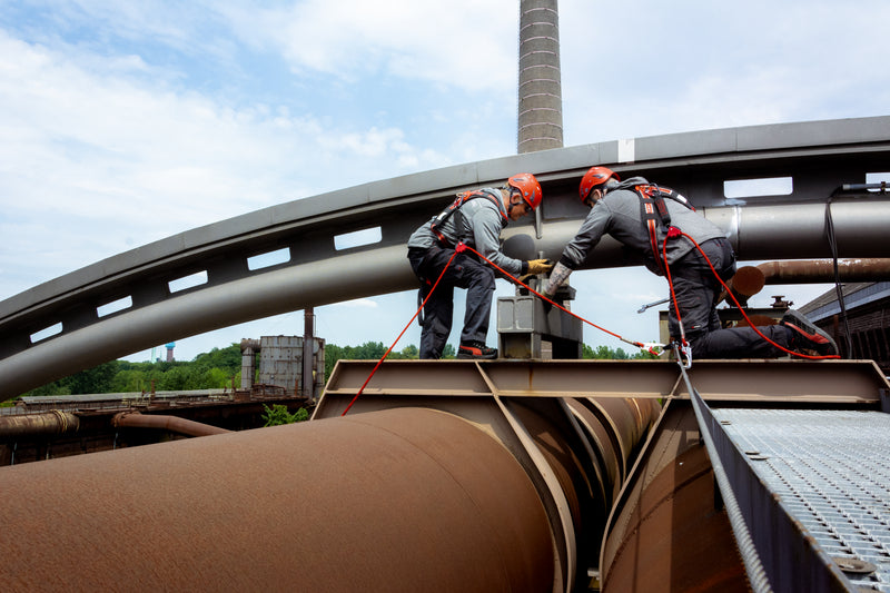 Zwei Arbeiter in voller Schutzausrüstung sichern sich mit Auffanggurten und Seilen auf einer Industrieanlage.