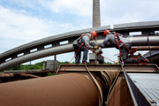 Zwei Arbeiter in voller Schutzausrüstung sichern sich mit Auffanggurten und Seilen auf einer Industrieanlage.