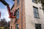 Ein Arbeiter in einer roten Hubarbeitsbühne reinigt Fenster an einer Backsteinfassade mit Auffanggurt und Helm.