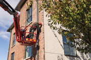 Ein Handwerker arbeitet gesichert mit Auffanggurt und Helm in einem Korb einer roten Hubarbeitsbühne an einer Fassade.
