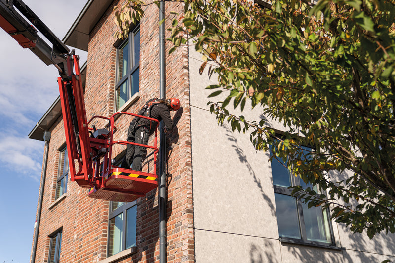 Ein Handwerker arbeitet gesichert mit Auffanggurt und Helm in einem Korb einer roten Hubarbeitsbühne an einer Fassade.