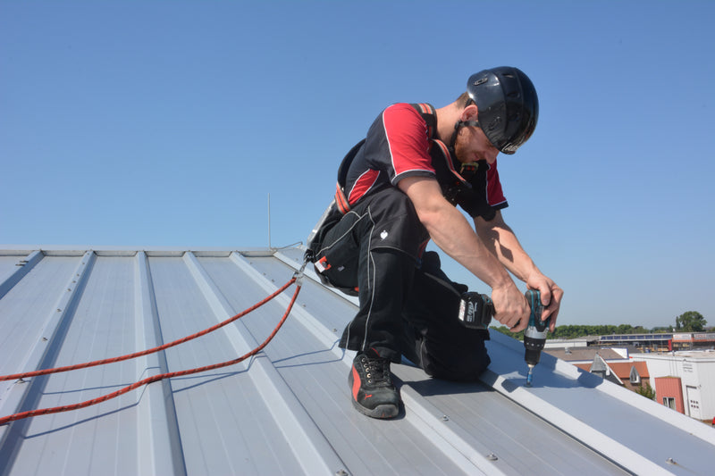 Ein Handwerker mit Schutzhelm und Auffanggurt arbeitet gesichert mit einem Akkuschrauber auf einem Metalldach.